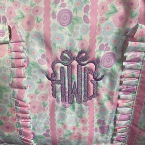 May include: Close-up of a floral patterned fabric in pink, white, and light blue. The design features roses and a monogram in a lavender color. Ruffled edges and vertical stripes add detail to the fabric.