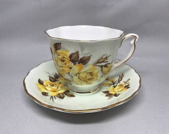 May include: A vintage teacup and saucer set. The cup and saucer are white with a pale green trim and feature a yellow rose floral design. The edges are trimmed with gold. A classic tea set.