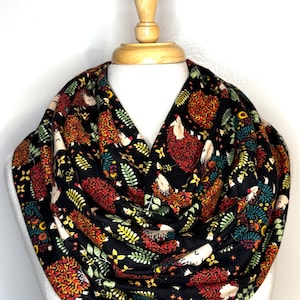 May include: Black infinity scarf with a repeating pattern of hedgehogs, flowers, and foliage in shades of red, orange, yellow, green, and white. The scarf is displayed on a mannequin with a wooden neck. The fabric appears soft and flowing.