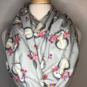 May include: A light blue infinity scarf featuring a repeating pattern of hedgehogs with floral bouquets. The scarf is wrapped around a mannequin, showcasing the design and style. The hedgehogs have white bellies, gray spines, and are surrounded by pink, red, and yellow flowers.
