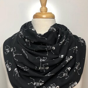May include: A black infinity scarf with a white skeleton pattern. The skeletons are dancing and have a skull for a head.