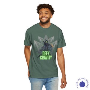 K&ouml;nnte beinhalten: Gr&uuml;nes T-Shirt mit einer Grafik einer Hexe mit einem Besen und Umhang, mit dem Text "DEFY GRAVITY" in Gr&uuml;n. Die Hexe ist vor einem wei&szlig;en, sternf&ouml;rmigen Hintergrund platziert. Das T-Shirt hat eine einfarbige gr&uuml;ne Farbe.