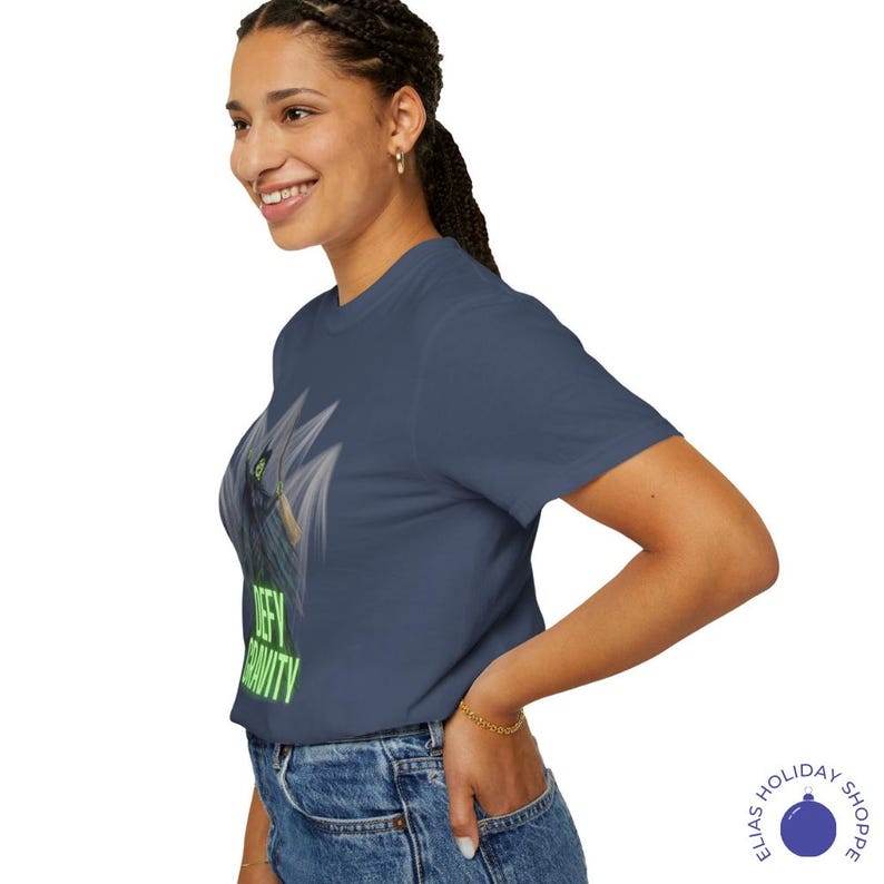 K&ouml;nnte beinhalten: Marineblaues T-Shirt mit einer Grafik einer gefl&uuml;gelten Figur und dem neongr&uuml;nen Text "Defy Gravity". Das Design befindet sich auf der Vorderseite des T-Shirts. Das T-Shirt besteht aus einem weichen Material.