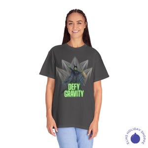 K&ouml;nnte beinhalten: Dunkelgraues T-Shirt mit einer Grafik einer gr&uuml;nen Hexe mit einem Besen, vor einem stilisierten Hintergrund. Die Worte "DEFY GRAVITY" sind in Neongr&uuml;n gehalten. Das Shirt ist ein bequemer, l&auml;ssiger Stil.