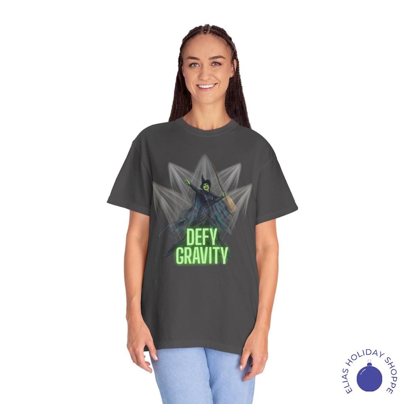 K&ouml;nnte beinhalten: Dunkelgraues T-Shirt mit einer Grafik einer gr&uuml;nen Hexe mit einem Besen, vor einem stilisierten Hintergrund. Die Worte "DEFY GRAVITY" sind in Neongr&uuml;n gehalten. Das Shirt ist ein bequemer, l&auml;ssiger Stil.