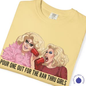 May include: Yellow Comfort Colors t-shirt featuring a graphic of two drag queens holding red cups. The text "POUR ONE OUT FOR THE RAN THRU GIRLS" is printed across the bottom of the image. The shirt has a crew neck.