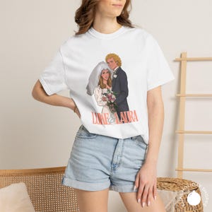 May include: White t-shirt featuring a cartoon illustration of a bride and groom, with the text "LUKE & LAURA" in pink. The bride wears a veil and holds a bouquet. The groom is in a black suit. The shirt is worn by a person.