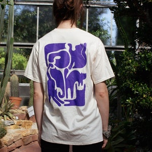 Puede incluir: Una persona lleva una camiseta blanca con un gráfico morado de varias formas abstractas en la espalda. La persona está de pie en un invernadero con cactus y otras plantas en el fondo.