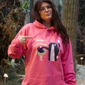 May include: A pink hoodie with a black cat graphic and the text "Stop Catcalling". The cat is holding a phone and the text is in a white font.