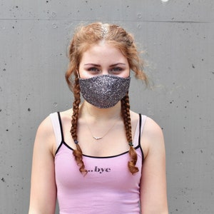 Könnte beinhalten: Eine junge Person mit langen braunen Haaren in Zöpfen trägt ein rosa Tanktop mit dem Aufdruck "...bye" auf der Vorderseite. Sie trägt eine glitzernde graue Gesichtsmaske.