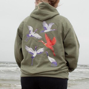 May include: A person wearing a green hooded sweatshirt with a design of white and red birds on the back. The person is standing on a beach with the ocean in the background.
