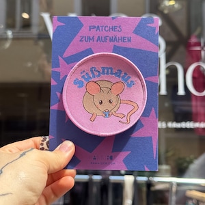 Può includere: Una toppa ricamata circolare raffigurante un topo marrone chiaro che tiene un oggetto blu, con la scritta "Süßmaus" in blu sopra. La toppa è montata su un cartoncino rettangolare viola con stelle blu e la scritta "PATCHES ZUM AUFNÄHEN".