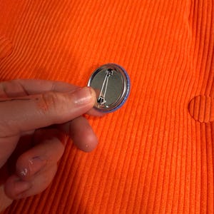 May include: A small, round button with a blue border and a shiny metal back. The button is held against a textured, orange corduroy fabric. The pin on the back is visible.