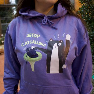 May include: Purple hoodie with a black and white cat illustration holding a phone and the text "STOP CATCALLING!" printed on the front.