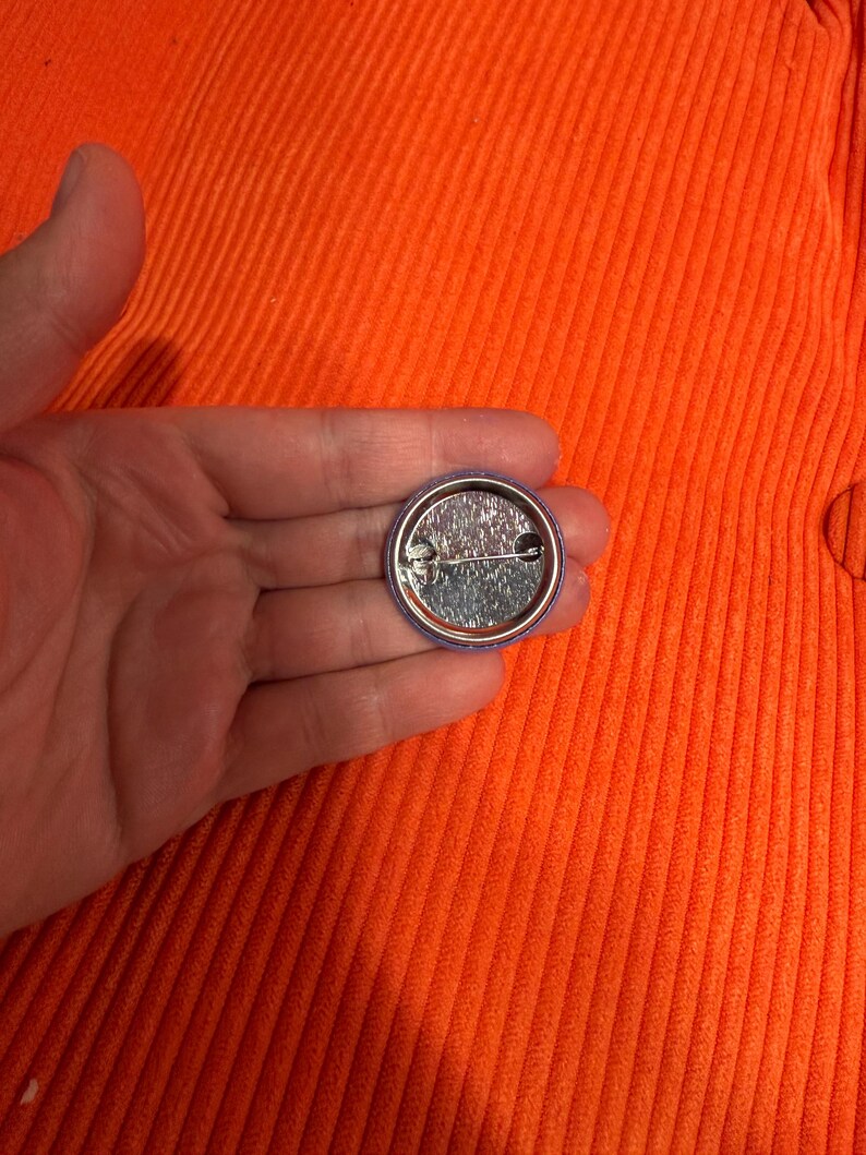 May include: A small, round button pin with a silver metal back and a safety pin clasp. The pin has a purple border and a shiny, reflective surface.
