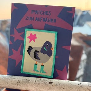 Könnte beinhalten: Ein rechteckiger, bestickter Aufnäher mit einem grauen Taubenmotiv, die türkisfarbene Gummistiefel und einen rosa Stern trägt, auf cremefarbenem Grund mit türkisfarbenem Rand. Der Aufnäher ist auf einer violetten Karte mit dem Text PATCHES ZUM AUFNÄHEN befestigt.