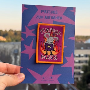 Op de afbeelding: Een rechthoekige stoffen patch met een oranje rand, voorzien van een getekende muis in een paarse cape voor oranje vlammen. De tekst luidt "FEMINIS" boven de muis en "MAUS" eronder, bevestigd op een paarse kaart met roze sterren.