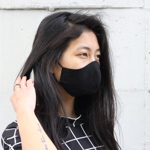 Può includere: Una persona che indossa una maschera facciale nera e una camicia a quadri neri e bianchi. La persona guarda a destra e ha i capelli lunghi e neri.