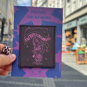 Puede incluir: Un parche rectangular de tela negra con una ilustración lineal rosa de un ratón sosteniendo una pancarta que dice ANTIFASCHISMAUS. El parche está montado sobre una tarjeta morada con patrones de estrellas y el texto PATCHES ZUM AUFNÄHEN y HANNISCH.COM.