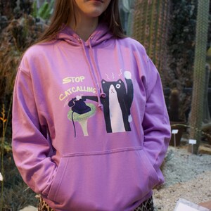 May include: A purple hoodie with a black cat holding a phone and the text "STOP CATCALLING" printed on the front.