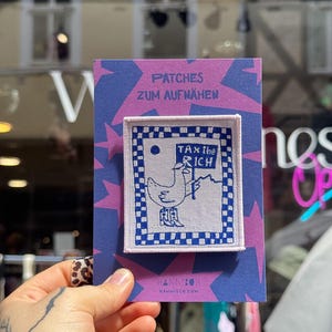 Peut inclure: Un écusson brodé rectangulaire représentant un oiseau blanc portant des bottes de cow-boy et tenant une pancarte avec l'inscription TAX THE RICH. L'écusson a une bordure à carreaux bleus et blancs sur une carte violette PATCHES ZUM AUFNÄHEN.
