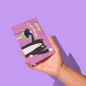 May include: A rectangular greeting card with a purple background is held in a hand. The card features a black and white cat cartoon with the text "STOP CATCALLING!" and a drawing of a telephone.