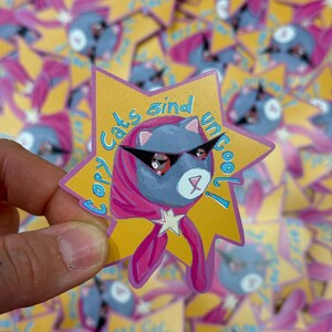 Könnte beinhalten: Ein sternförmiger Aufkleber mit einer Cartoon-Katze, die eine Sonnenbrille und einen rosa Schal trägt. Die Katze ist blau mit einem weißen Stern auf der Brust. Der Aufkleber hat den Text "Copy cats sind uncool!" in Englisch und Deutsch.