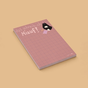 May include: A rectangular notepad with a dusty rose grid pattern. The top left features the text "das schreib ich Miauf!" in white. A black cat illustration with a white star on its chest holds a pencil next to a small pink note.