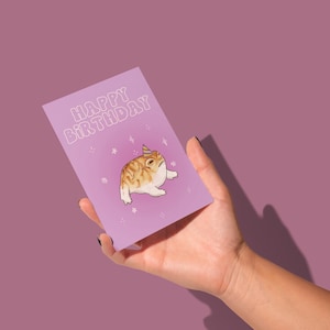 Könnte beinhalten: Eine lila Geburtstagskarte mit dem Text "HAPPY BIRTHDAY" in Weiß, die einen Frosch mit Partyhut zeigt. Der Frosch ist braun und weiß mit einem mürrischen Gesichtsausdruck. Die Karte wird in einer Hand vor einem lila Hintergrund gehalten.