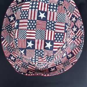 May include: A bucket hat featuring a patchwork pattern of red and white stripes and blue squares with white stars. The design is arranged in a grid across the crown and the wide, downward-sloping brim, finished with a thin red trim along the edge.