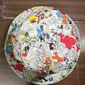 May include: A bucket hat featuring a white background with a dense, colorful all-over print of various Looney Tunes cartoon characters including Bugs Bunny, Daffy Duck, Porky Pig, Taz, Sylvester, and Marvin the Martian in red, yellow, green, and blue.