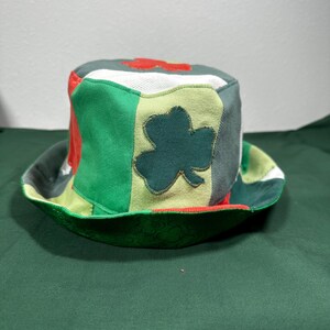 May include: A patchwork bucket hat with a St. Patrick's Day theme. The hat features green, red, and white fabric panels, with a large green shamrock applique on the side. The brim is green with a subtle pattern.