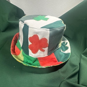 May include: A patchwork top hat with a wide brim, featuring a mix of green, white, and red fabrics. The hat is adorned with green and red shamrock appliqués, perfect for St. Patrick's Day celebrations.