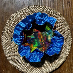 May include: A round woven basket with a blue satin drawstring bag. The bag's interior features a colorful abstract pattern with red, yellow, green, and purple hues. The bag is sitting on a round woven basket.