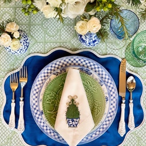 May include: A white linen napkin with an embroidered green Christmas tree in a blue and white pot. The napkin is folded and placed on a blue and white patterned plate, which is on a green and white patterned placemat.