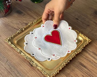 Peut inclure: Deux dessous-de-verre en lin blanc avec des bords festonnés et des cœurs brodés rouges. Les dessous-de-verre sont sur un plateau doré.