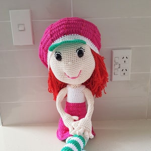 Könnte beinhalten: Eine handgefertigte Häkelpuppe mit weißem Gesicht, rotem Haar und einem rosa Hut mit grünem Visier. Die Puppe trägt ein weißes Oberteil, einen rosa Rock und grün-weiß gestreifte Beine. Die Füße der Puppe sind rosa.