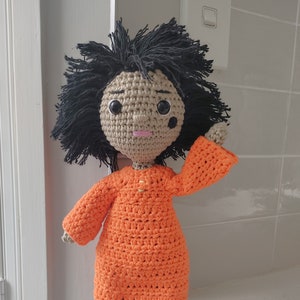 Puede incluir: Una muñeca de crochet con pelo negro, piel morena y boca rosa. La muñeca lleva un vestido naranja.