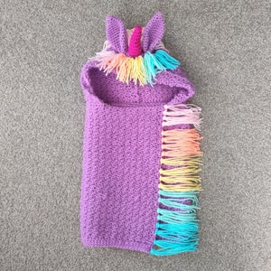 May include: A purple crocheted unicorn hooded blanket. The hood features purple ears, a pink horn, and a rainbow mane. The blanket has a fringe of rainbow-coloured yarn along the side.