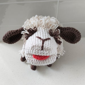 Puede incluir: Un juguete de oveja de crochet blanco con orejas marrones y una boca roja. La oveja tiene un cuerpo blanco esponjoso y una nariz marrón.