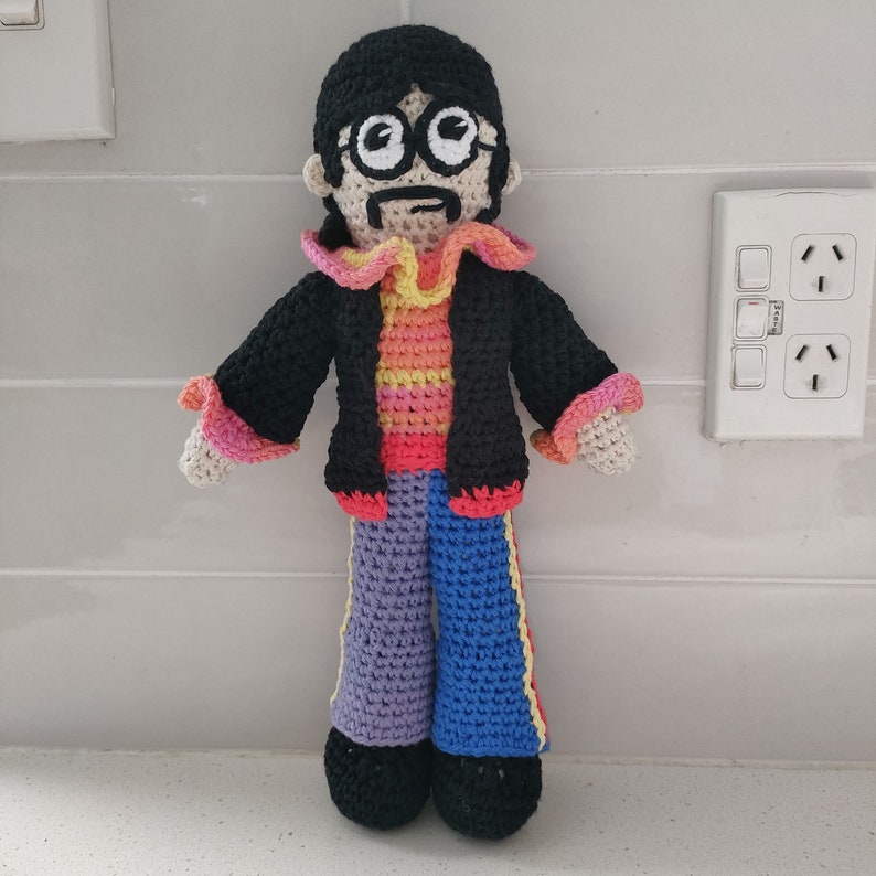 May include: A crocheted doll with black hair, a black jacket, a rainbow striped shirt, and blue and purple pants. The doll has a mustache and wears round glasses.