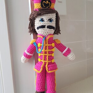 May include: A crocheted doll wearing a pink hat with a yellow band and a black emblem. The doll is wearing a pink and yellow striped uniform with a blue lanyard. The doll has brown hair, white skin, blue eyes, and a black mustache.