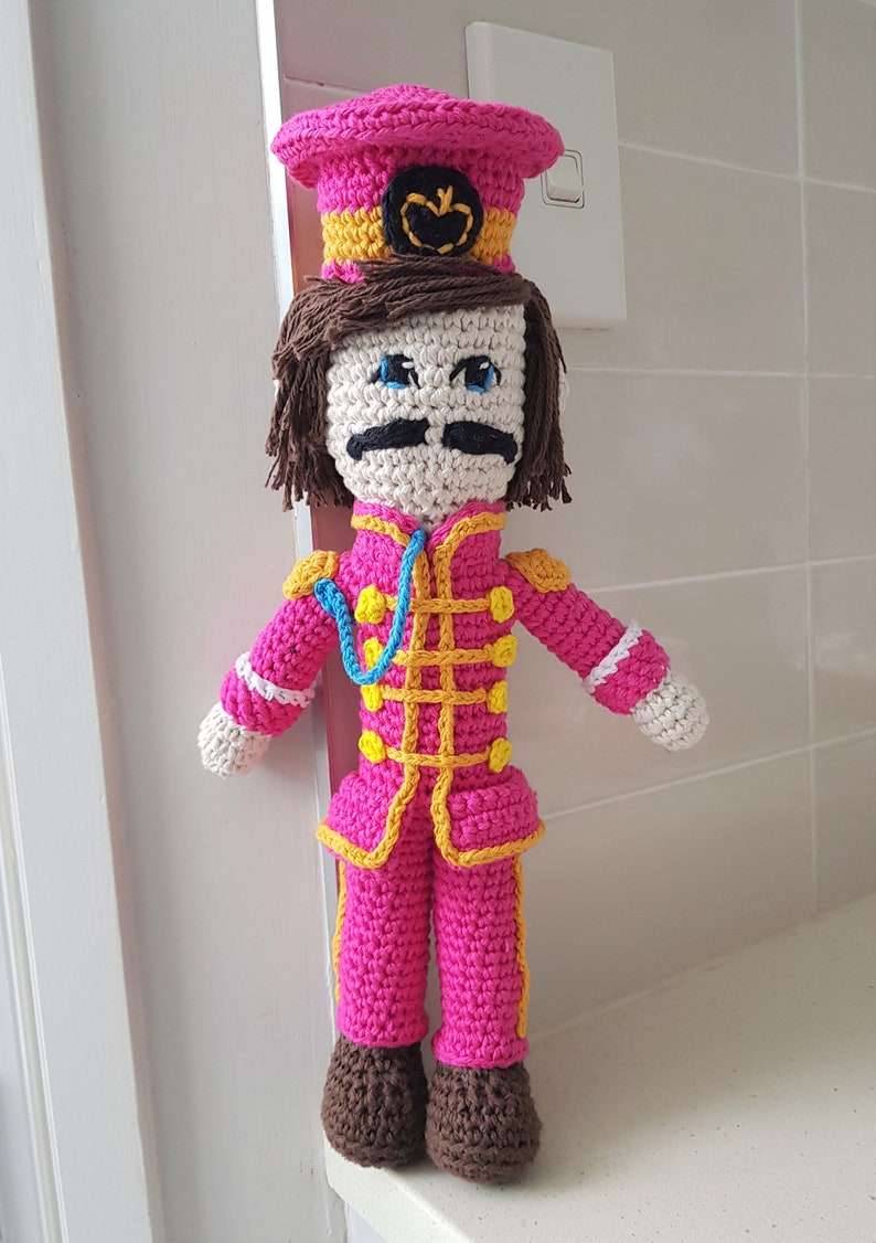 May include: A crocheted doll wearing a pink hat with a yellow band and a black emblem. The doll is wearing a pink and yellow striped uniform with a blue lanyard. The doll has brown hair, white skin, blue eyes, and a black mustache.