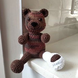 Può includere: Un orsacchiotto all'uncinetto fatto a mano nei toni del marrone, rosso e beige. L'orso ha naso e occhi neri. Una gamba è ingessata di bianco. L'orso è seduto su una superficie bianca.