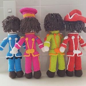 May include: Four crocheted dolls dressed in colorful uniforms with hats. The dolls are standing in a row on a white tiled surface.
