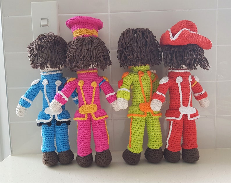 May include: Four crocheted dolls dressed in colorful uniforms with hats. The dolls are standing in a row on a white tiled surface.