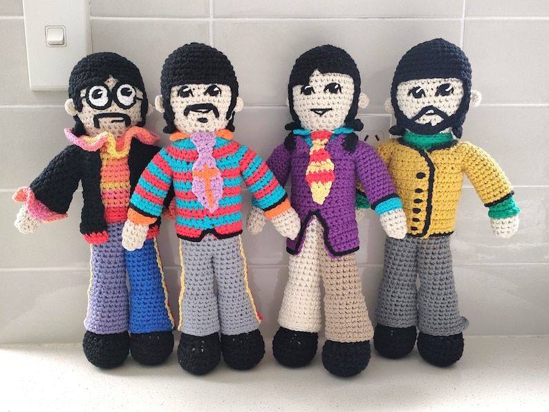 May include: Four crocheted dolls dressed as the Beatles. The dolls are wearing colorful clothing and have black hair. The dolls are standing on a white tiled surface.
