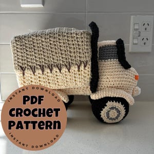 May include: A crocheted toy truck in beige, tan, and black. The truck features a detailed design with a cab, wheels, and a cargo bed. The text "PDF CROCHET PATTERN" is displayed on a circular graphic in the lower left corner.