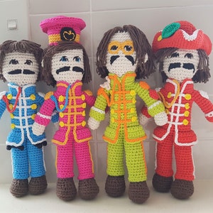 May include: Four crocheted dolls dressed in colorful outfits, resembling the Beatles. Each doll has a unique hairstyle and facial features, with brown hair, mustaches, and glasses. The dolls are standing on a white surface.
