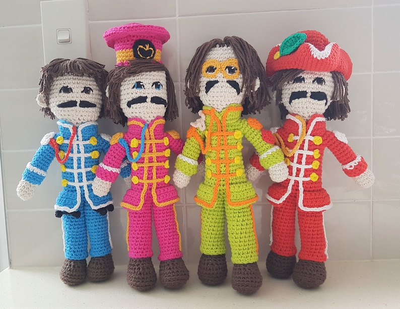 May include: Four crocheted dolls dressed in colorful outfits, resembling the Beatles. Each doll has a unique hairstyle and facial features, with brown hair, mustaches, and glasses. The dolls are standing on a white surface.